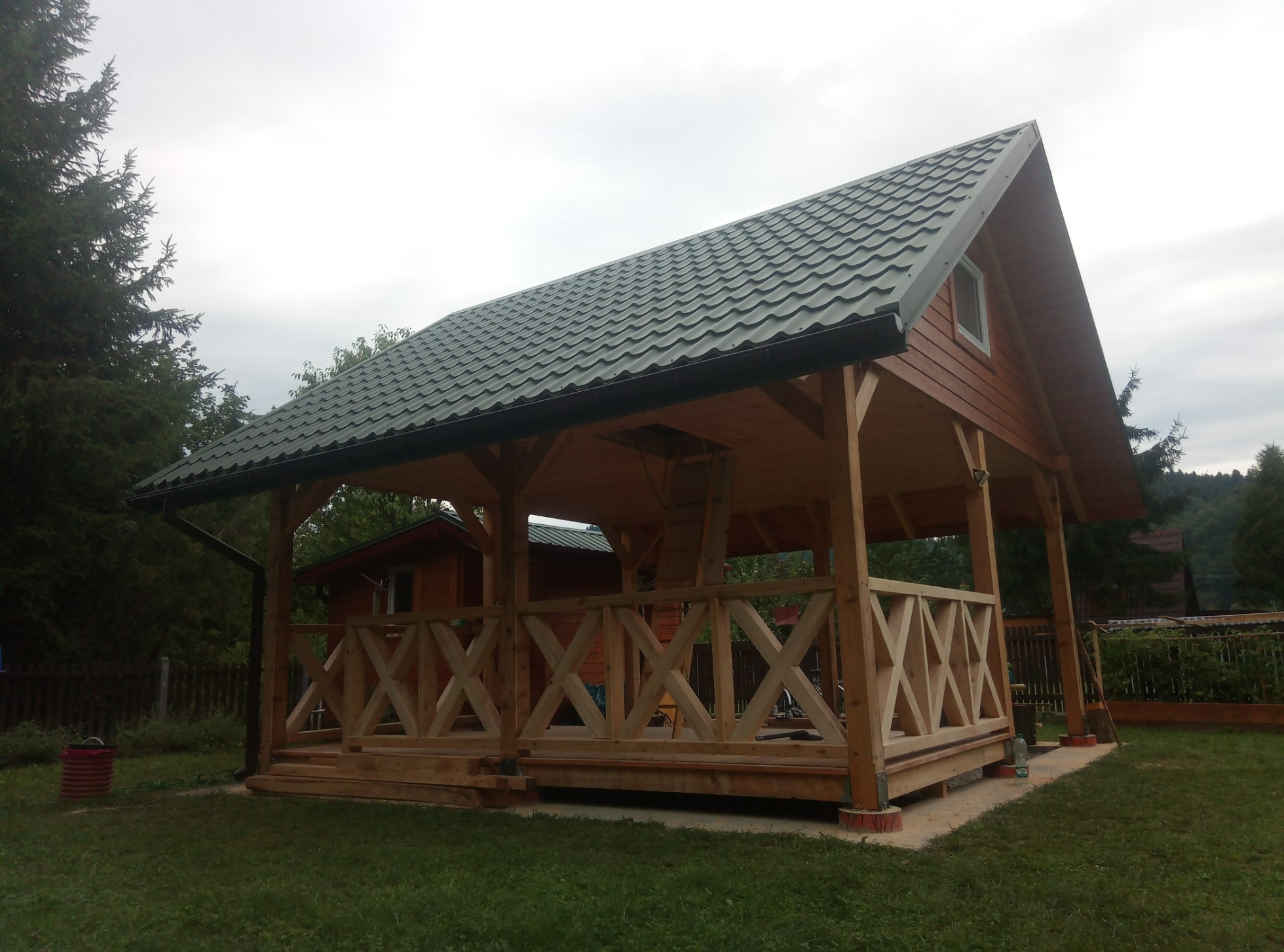 Drewniana wiata garażowa z zielonym dachem z blachodachówki, kratowane balustrady, widok na ogród i mały domek w tle.