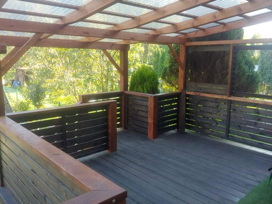 Zadaszona drewniana altana z czarną podłogą i balustradą, otoczona zielenią, z widocznym zadaszeniem z przezroczystych płyt.