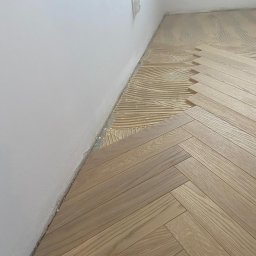 Firma Gawin - Układanie parkietu w jodełkę: widoczny fragment podłogi z klejem i ułożonymi deskami, przy białej ścianie z gniazdkiem elektrycznym.