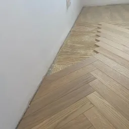 Układanie parkietu w jodełkę: widoczny fragment podłogi z klejem i ułożonymi deskami, przy białej ścianie z gniazdkiem elektrycznym.