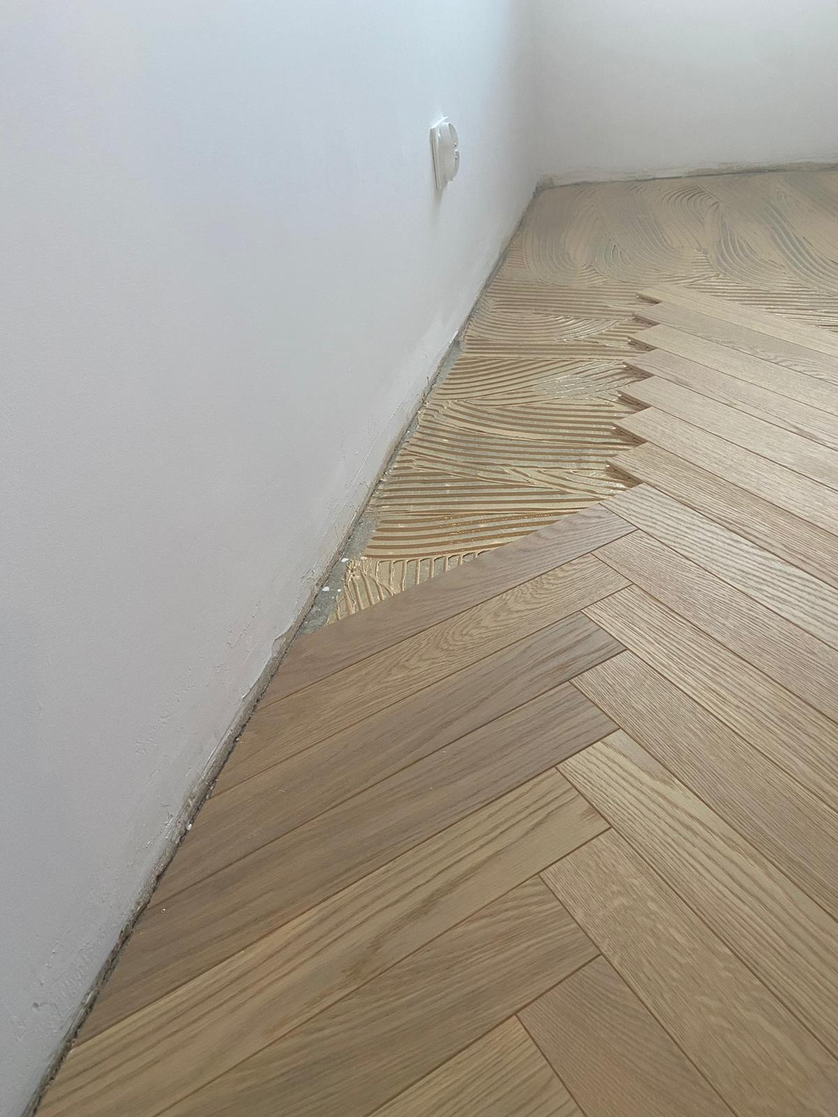 Układanie parkietu w jodełkę: widoczny fragment podłogi z klejem i ułożonymi deskami, przy białej ścianie z gniazdkiem elektrycznym.