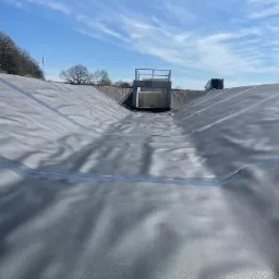 Wyłożony geomembraną zbiornik retencyjny z betonową konstrukcją w tle, widoczny samochód dostawczy i błękitne niebo z chmurami.