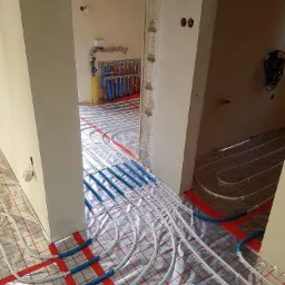 Widok na rozłożoną instalację ogrzewania podłogowego z niebieskimi rurami na folii aluminiowej z czerwoną siatką, widoczne rozdzielacze i nieotynkowane ściany w tle.