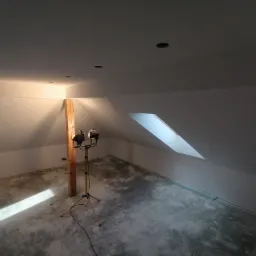 Poddasze po nałożeniu gładzi, oświetlone lampą budowlaną na statywie, widoczne przygotowania do dalszych prac wykończeniowych.