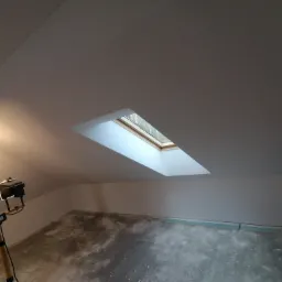 Poddasze po szpachlowaniu, widoczne okno dachowe, lampa budowlana i surowa podłoga przygotowana do dalszych prac wykończeniowych.