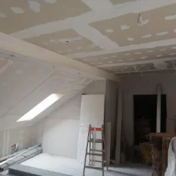 Poddasze w trakcie prac wykończeniowych: widoczne płyty karton-gips na skosach i suficie, częściowo zaszpachlowane, z widocznymi przewodami elektrycznymi i oknem dachowym. W tle oparta o ścianę...