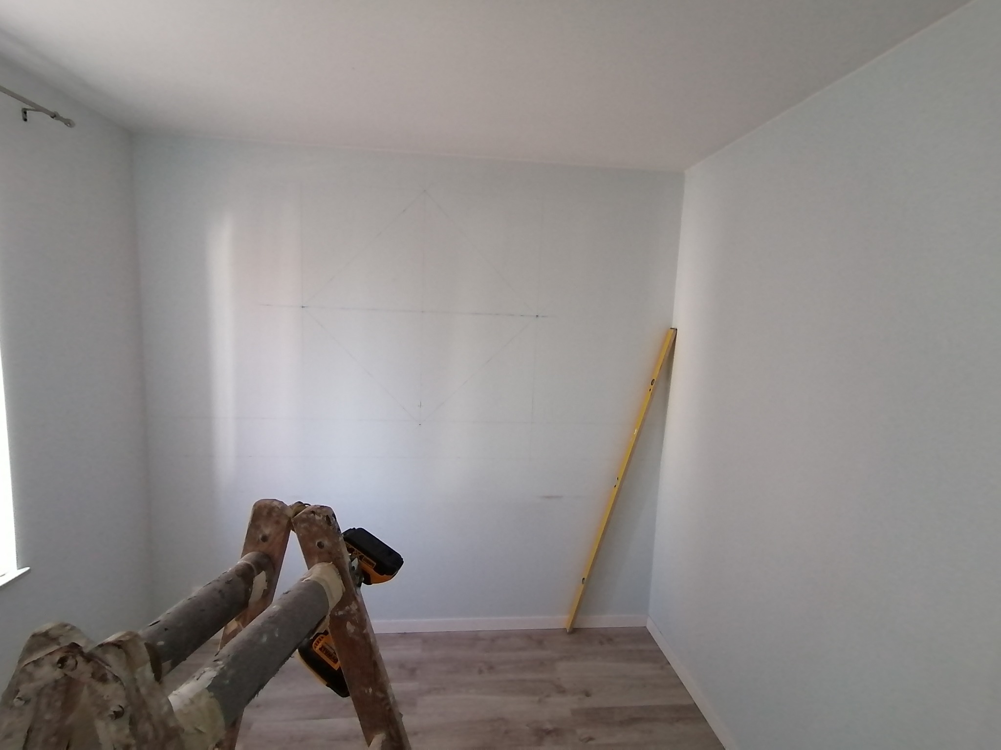 Pokój w trakcie przygotowania do prac wykończeniowych, widoczne ślady ołówka na ścianie wyznaczające wzór, drabina malarska z wkrętarką i poziomica oparta o ścianę.