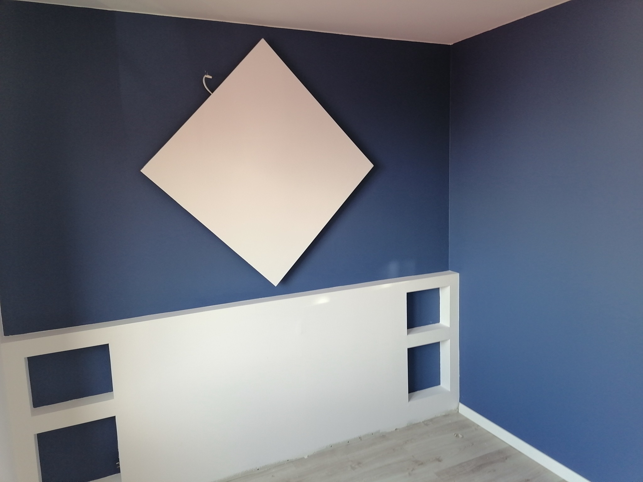 Niebieska ściana z białym, kwadratowym panelem akustycznym zamontowanym na skos, poniżej biała konstrukcja z półkami.