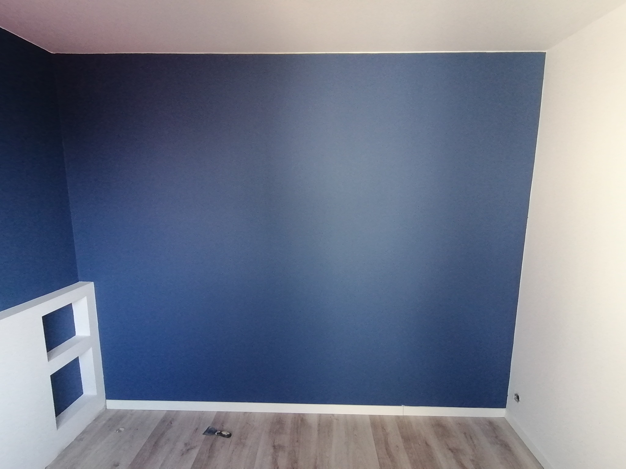 Pomieszczenie po malowaniu ścian na ciemny odcień niebieskiego, z widocznymi panelami podłogowymi i fragmentem białej konstrukcji z otworami.