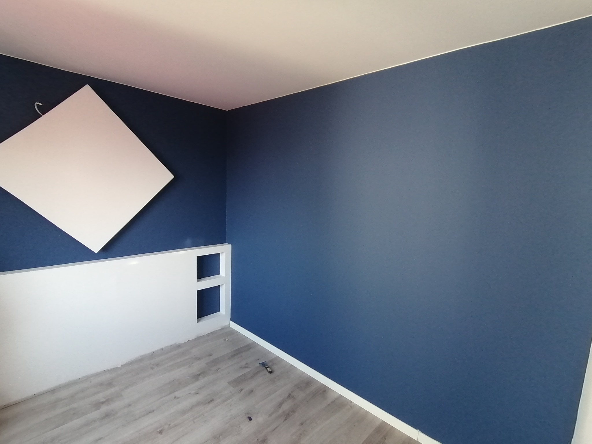 Pokój po świeżym malowaniu na granatowo, z białym panelem dekoracyjnym przy podłodze i kwadratowym panelem akustycznym na ścianie, widoczne panele podłogowe.