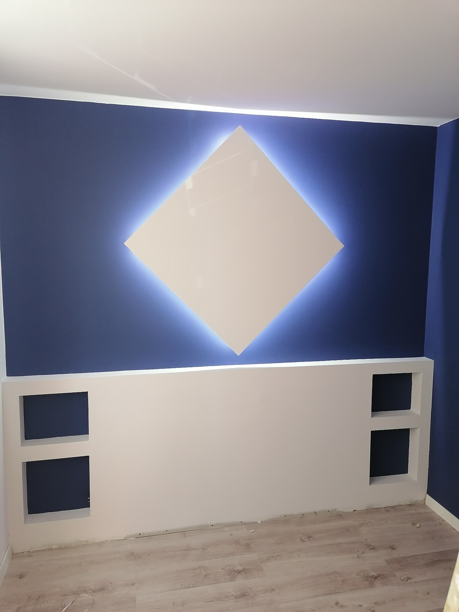 Niebieska ściana z podświetlanym rombem i białą, niską zabudową z wnękami, jasna podłoga.
