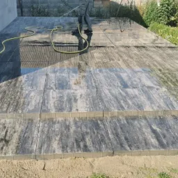 Układanie kostki brukowej: Czyszczenie wodą nowo ułożonej, szarej kostki brukowej z delikatnym wzorem drewna, z widocznymi dwoma stopniami i żółtym wężem ogrodowym.