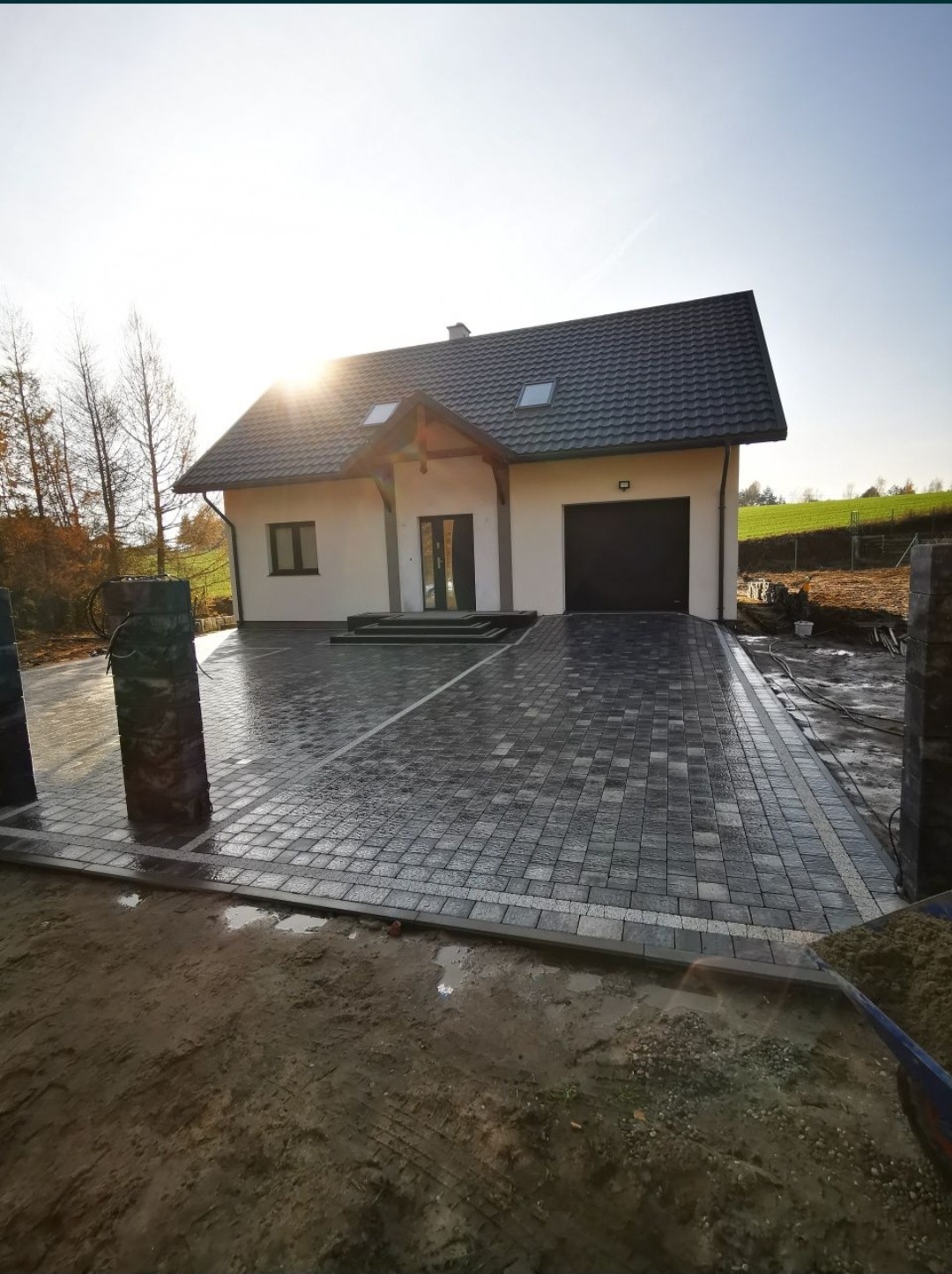 Nowo ułożona, mokra kostka brukowa w odcieniach szarości prowadząca do domu z garażem; widoczne słupki ogrodzeniowe i wózek z piaskiem.