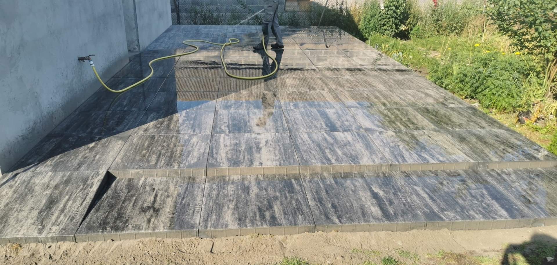 Układanie kostki brukowej: Czyszczenie wodą nowo ułożonej, szarej kostki brukowej z delikatnym wzorem drewna, z widocznymi dwoma stopniami i żółtym wężem ogrodowym.