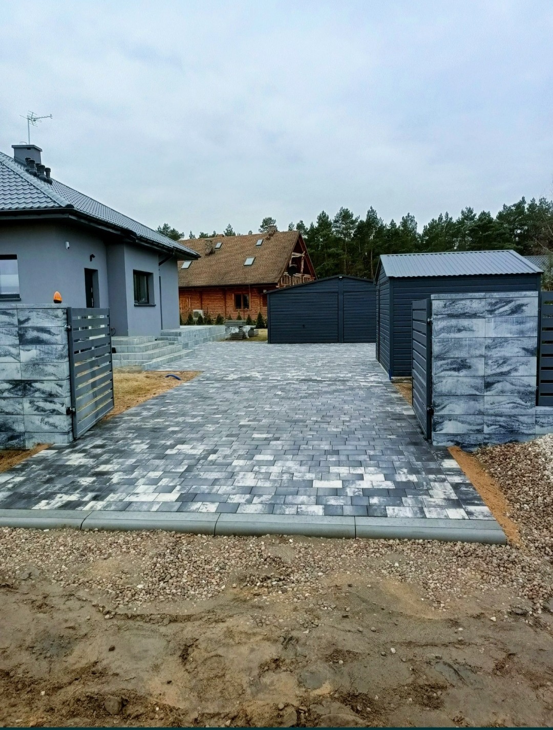 Ułożona kostka brukowa w odcieniach szarości prowadząca do domu z szarą elewacją i metalowego garażu, widoczne obrzeża i fragmenty gruntu z kamieniami.