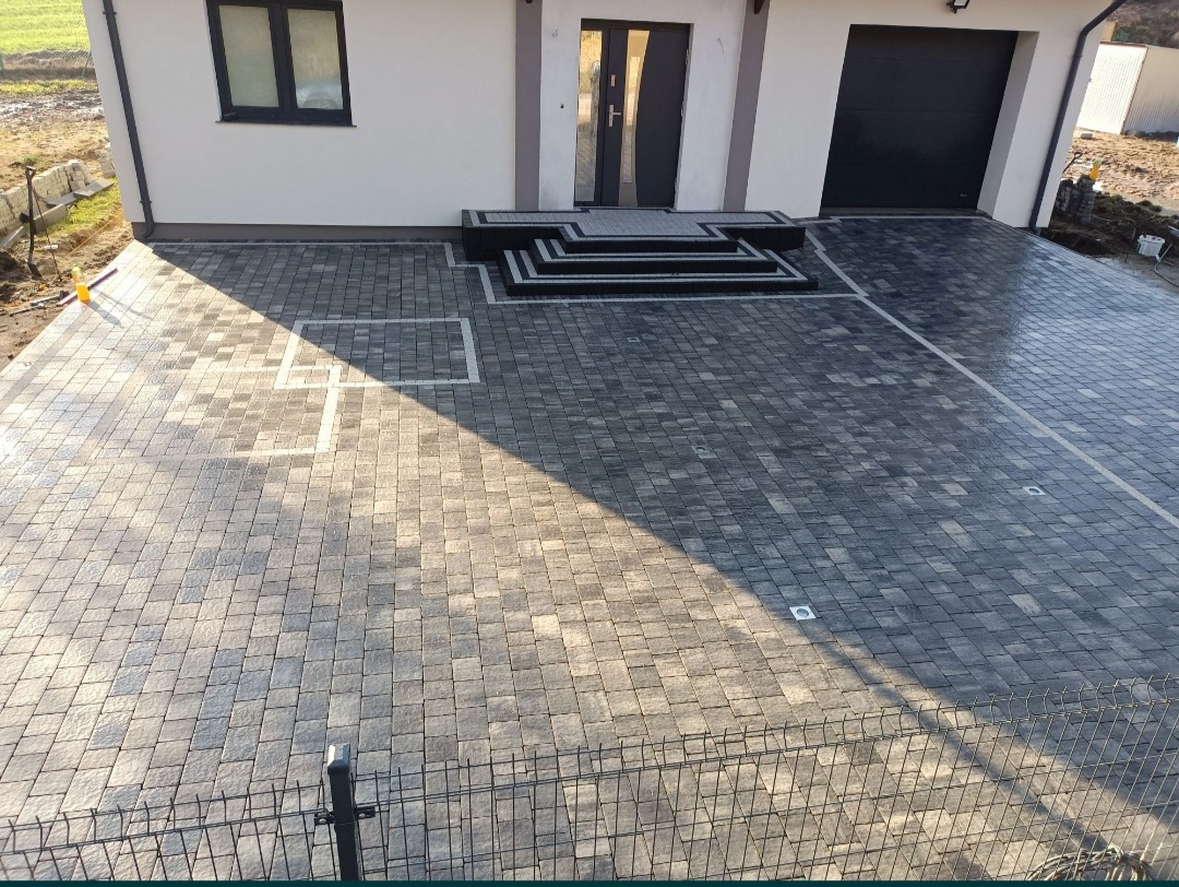 Nowoczesny podjazd z kostki brukowej w odcieniach szarości, z białymi liniami wyznaczającymi strefy, prowadzący do domu z garażem i schodami wejściowymi, widoczny fragment ogrodzenia.