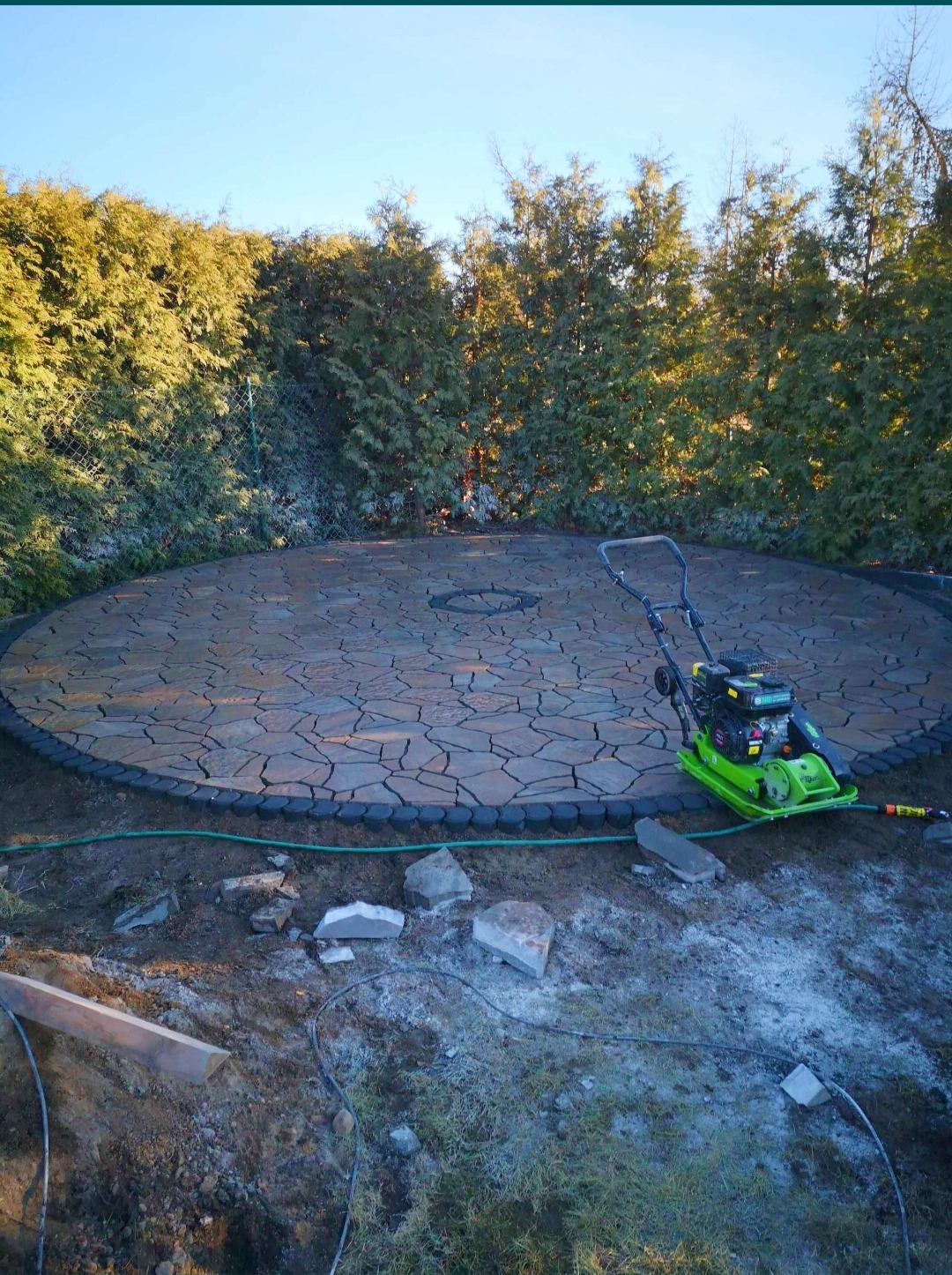 Okrągły plac z nieregularnej kostki brukowej otoczony ciemną obwódką, z zieloną zagęszczarką w tle żywopłot z iglaków.