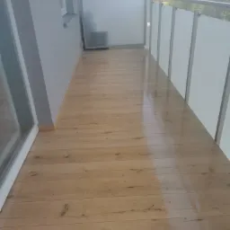 Jasnobrązowa podłoga z paneli imitujących drewno na balkonie z białymi panelami osłonowymi i szarą ścianą, widok na drzewa w tle.