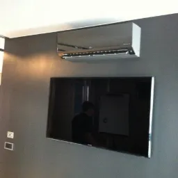 Srebrny klimatyzator ścienny LG zamontowany nad wiszącym telewizorem na szarej ścianie. Widoczne gniazdka elektryczne po lewej stronie.