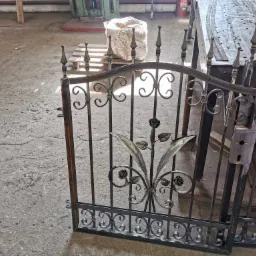 Fragment metalowej bramy z dekoracyjnymi elementami roślinnymi w warsztacie, widoczne spawane łączenia, surowy beton na podłodze, w tle materiały i narzędzia.