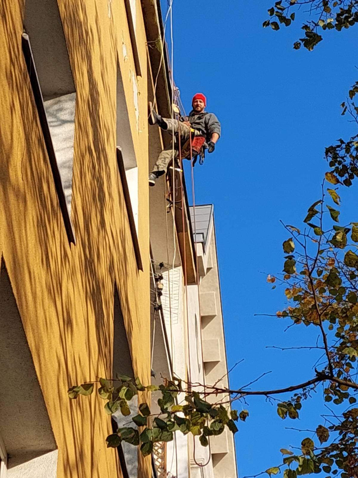 Pracownik na linach, w czerwonej czapce, w trakcie pracy na wysokości na żółtej elewacji budynku, widok z dołu, błękitne niebo i gałęzie drzew w tle.