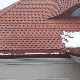 Czerwona dachówka karpiówka na dachu domu z oknem w kształcie półkola i śniegiem przy krawędziach.