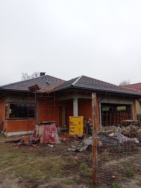 Dom jednorodzinny w trakcie budowy, z częściowo pokrytym dachem czerwoną dachówką ceramiczną, widoczne rusztowanie na dachu, cegły i materiały budowlane na ziemi, tablica informacyjna i ogrodzenie...