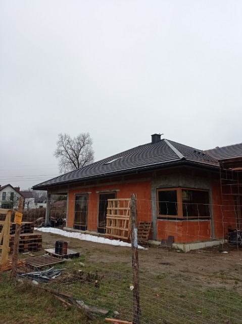Dom jednorodzinny w trakcie budowy z nowym, ciemnoszarym dachem, widoczne palety i ogrodzenie z siatki, pochmurne niebo w tle.