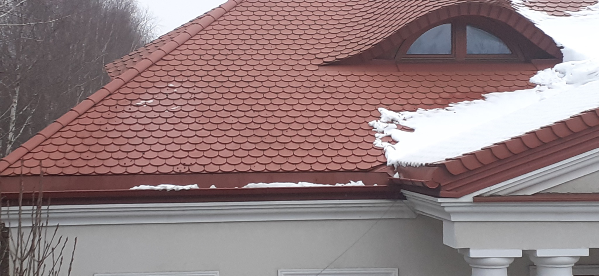 Czerwona dachówka karpiówka na dachu domu z oknem w kształcie półkola i śniegiem przy krawędziach.