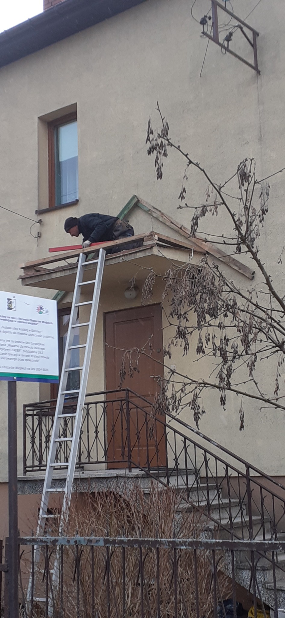 Pracownik na drabinie poziomuje łatę na zadaszeniu wejścia do budynku mieszkalnego, widoczny fragment dachu, drzwi wejściowe i tablica informacyjna.