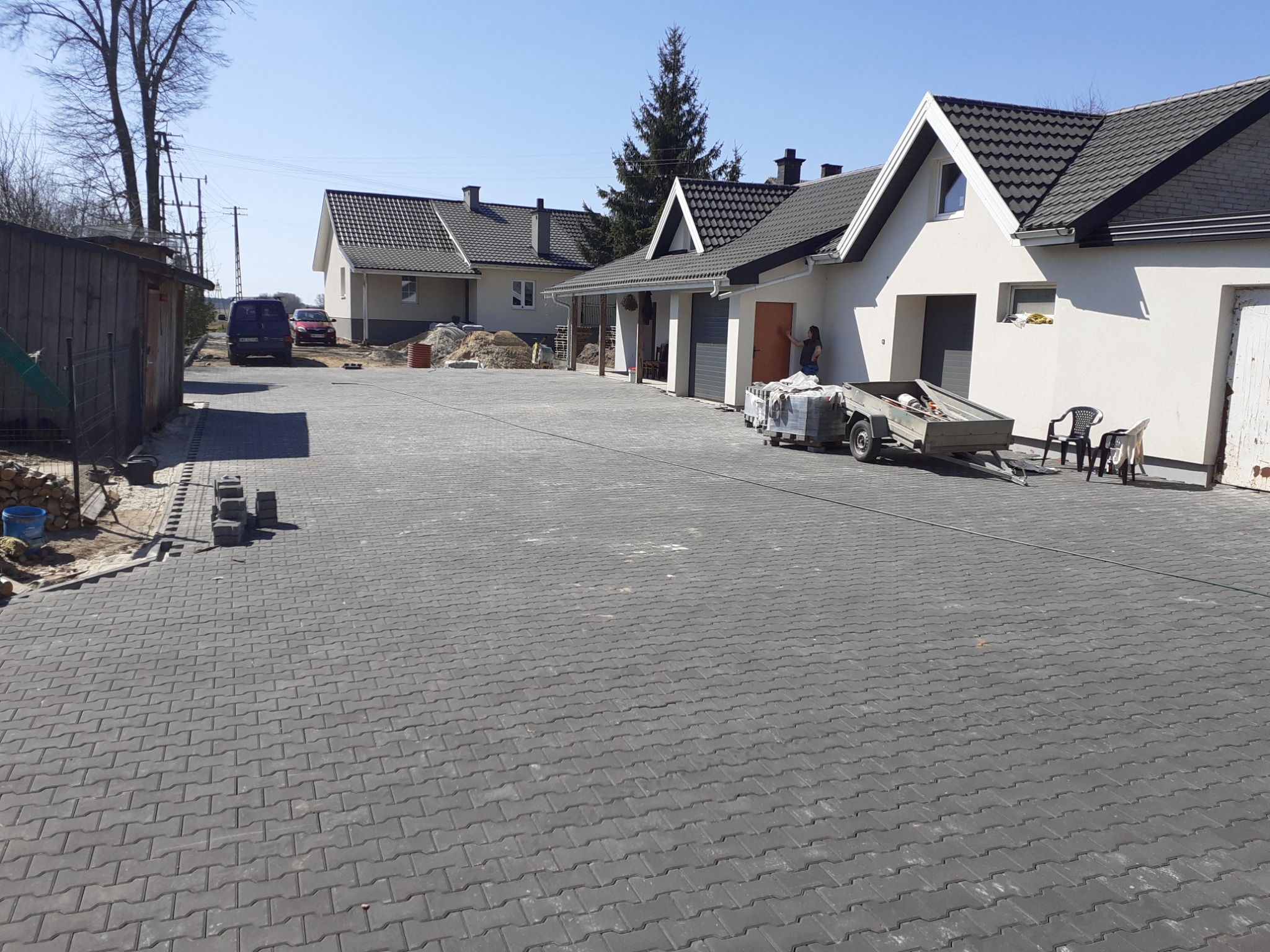 Nowe, grafitowe pokrycie dachowe na domach jednorodzinnych, widoczne rynny, kostka brukowa na podwórzu, przygotowany przyczepka z materiałami budowlanymi.