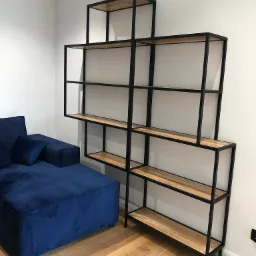 Narożnik obity granatowym welurem zestawiony z asymetrycznym regałem z drewnianymi półkami i czarną, metalową ramą na drewnianej podłodze.