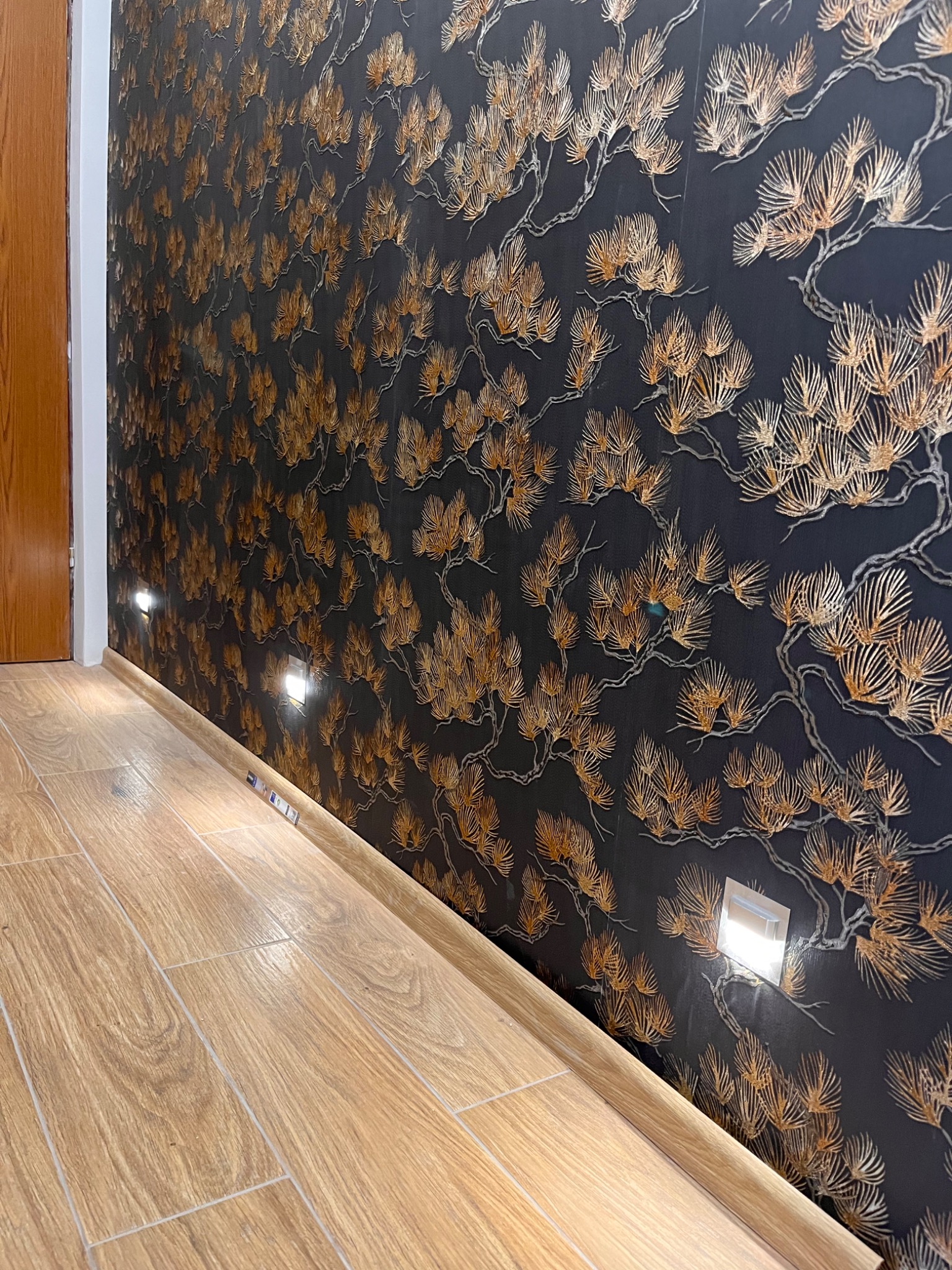 Fragment wykończonego wnętrza z drewnopodobną podłogą, czarną tapetą w złote motywy roślinne oraz wbudowanymi w ścianę punktowymi lampkami LED.