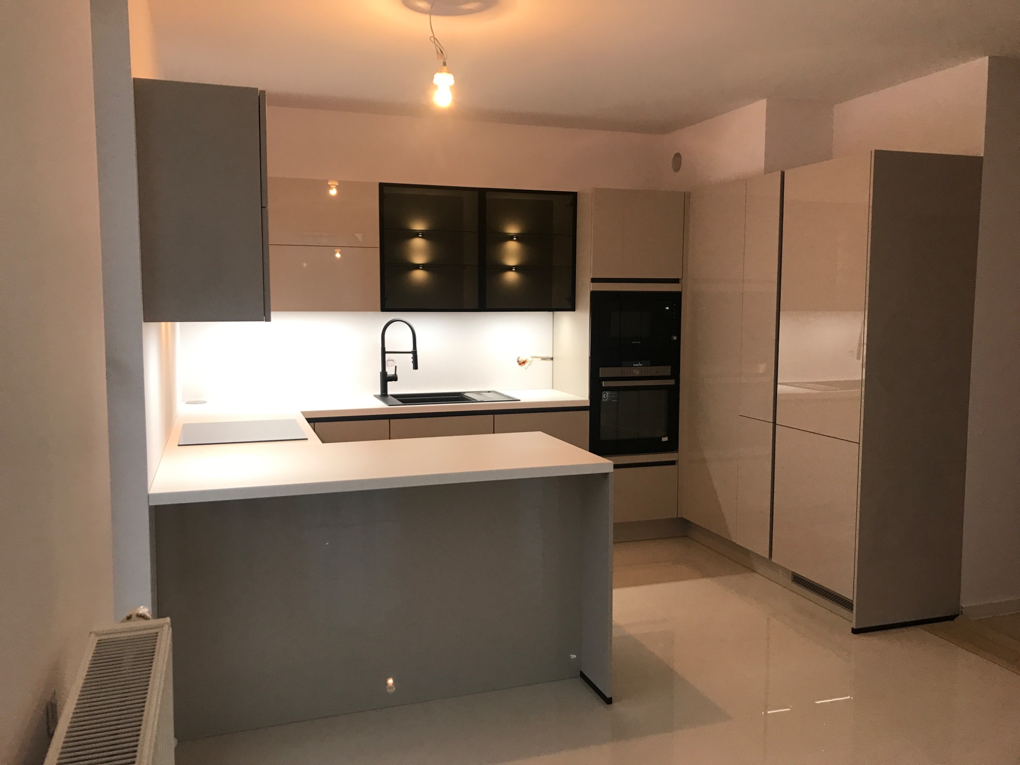 Nowoczesna, jasna kuchnia z wyspą, fronty szafek lakierowane na wysoki połysk, czarny zlew i bateria, podświetlane szafki wiszące z czarnymi ramami, widoczny grzejnik.