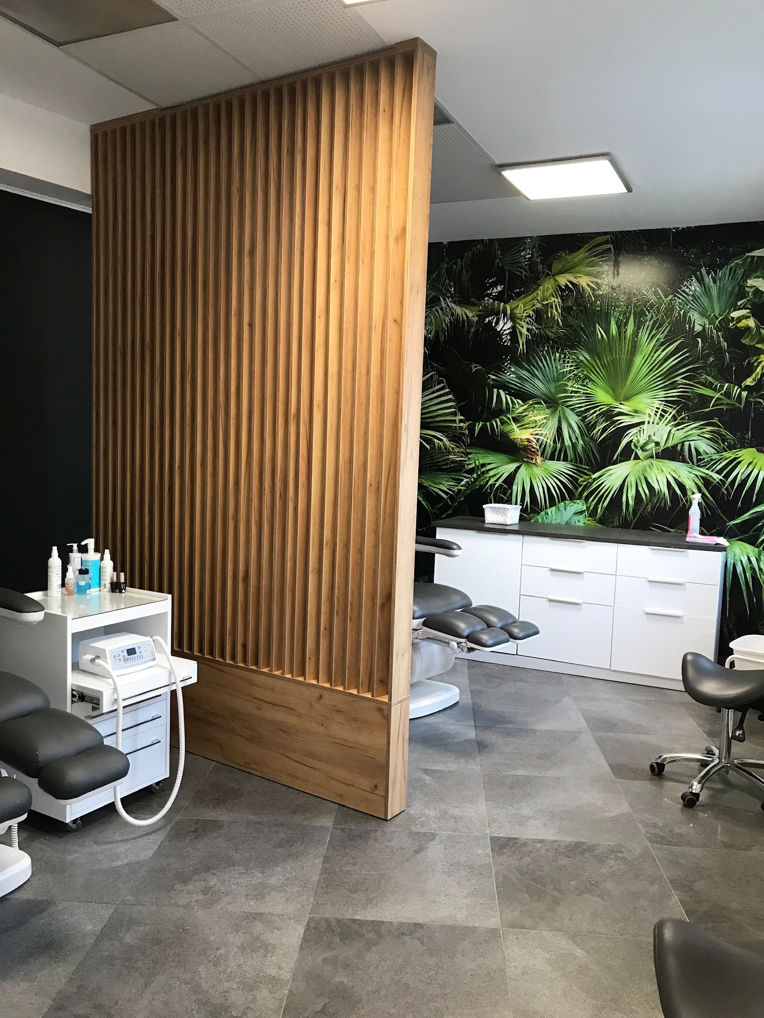 Nowoczesne wnętrze gabinetu kosmetycznego z drewnianą ażurową ścianką działową, fototapetą z motywem roślinnym, szarymi płytkami podłogowymi i białymi szafkami oraz dwoma fotelami zabiegowymi.
