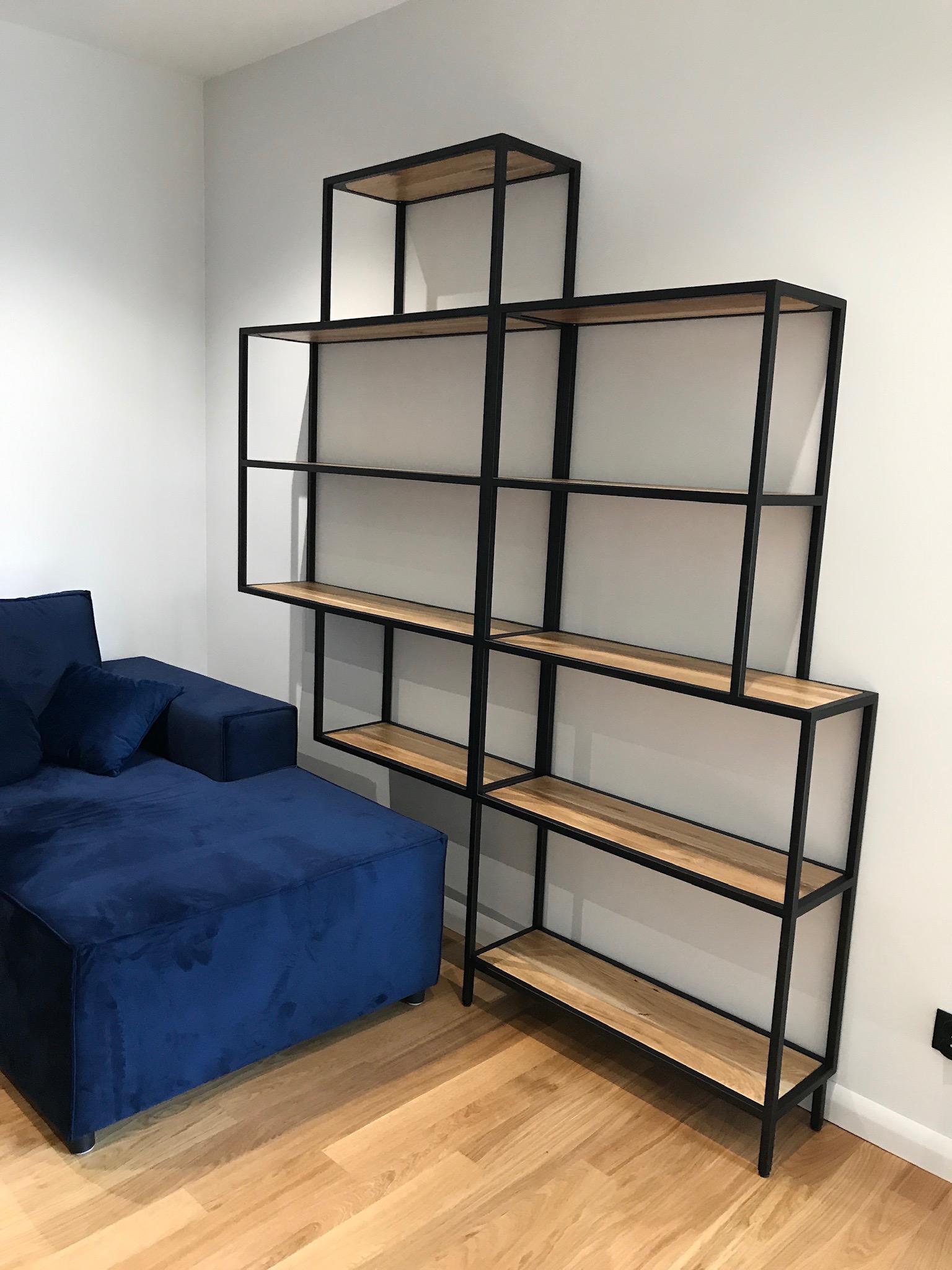 Narożnik obity granatowym welurem zestawiony z asymetrycznym regałem z drewnianymi półkami i czarną, metalową ramą na drewnianej podłodze.