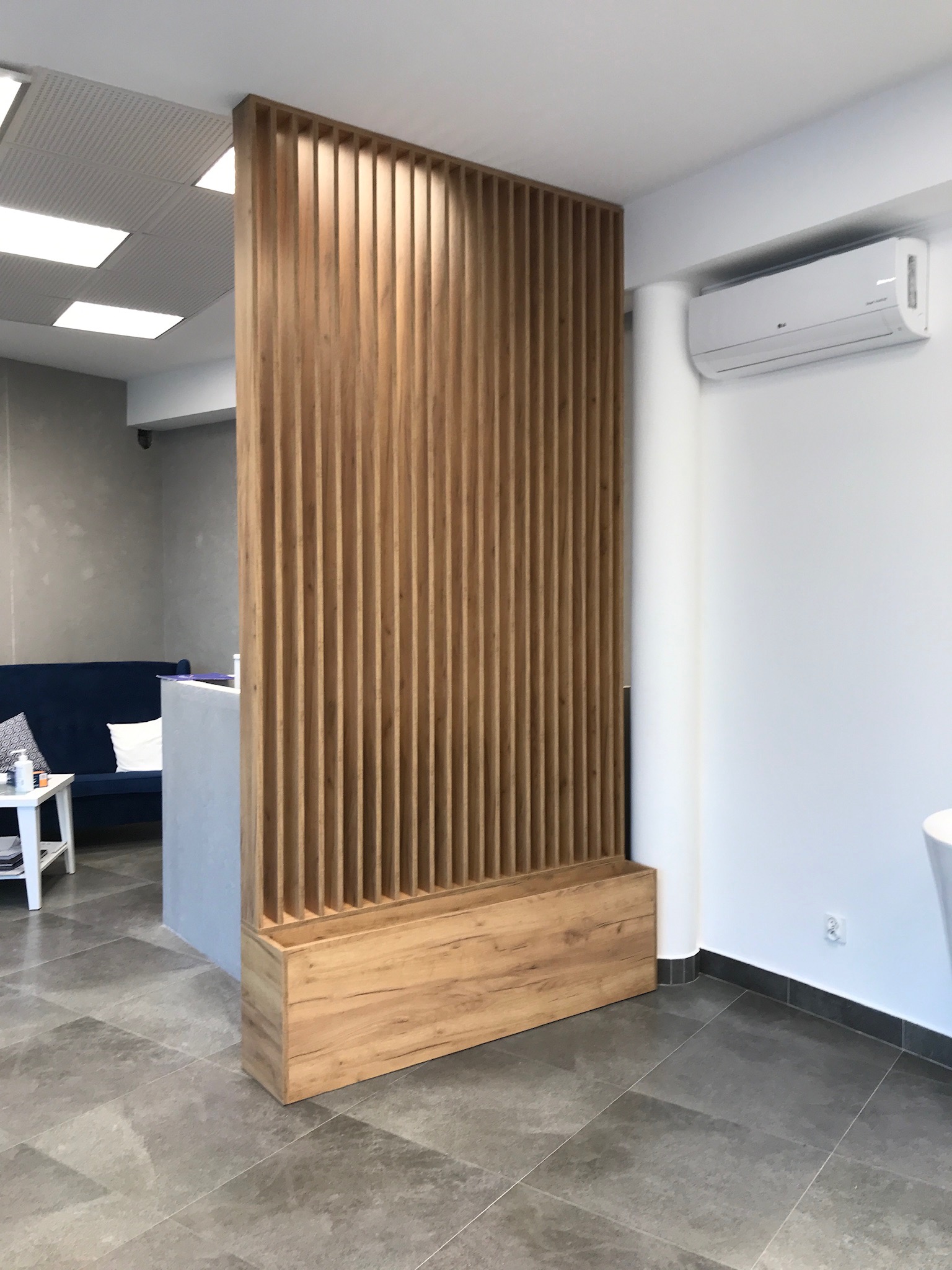 Drewniana ścianka działowa z pionowych listew, oddzielająca przestrzeń biurową z recepcją i strefą oczekiwania, widoczna klimatyzacja na białej ścianie.