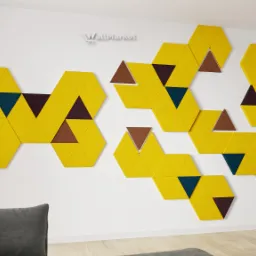 Żółte, szare i granatowe panele akustyczne w kształcie heksagonów i trójkątów, tworzące dekoracyjną kompozycję na białej ścianie w salonie z szarą sofą i fotelem.
