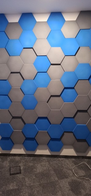 Ściana biura pokryta akustycznymi panelami ściennymi 3D w kształcie heksagonów w odcieniach szarości i błękitu, widoczny fragment szarej wykładziny podłogowej.