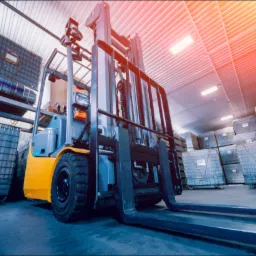 Żółto-szary wózek widłowy podnosi metalową skrzynię z zawartością w magazynie z rzędami podobnych skrzyń, oświetlonymi światłem z góry.