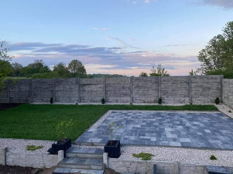 Nowoczesna aranżacja ogrodu z szarej kostki brukowej, trawnika i betonowego ogrodzenia na tle pastelowego nieba o zmierzchu.