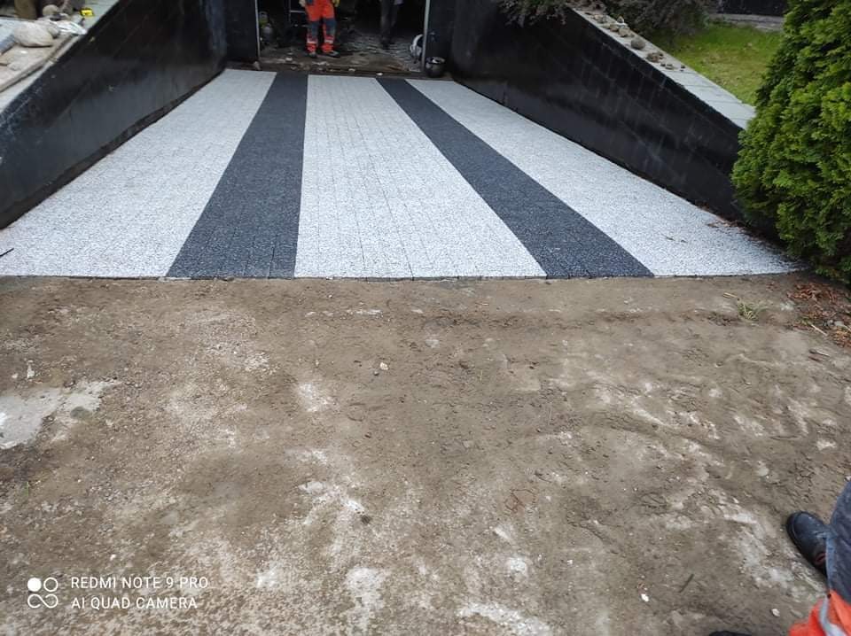 Podjazd do garażu z ułożoną dwukolorową kostką, jasnoszarą i grafitową, w pasy. W tle widoczny garaż oraz osoby w strojach roboczych. Na pierwszym planie teren przygotowany pod dalsze prace.
