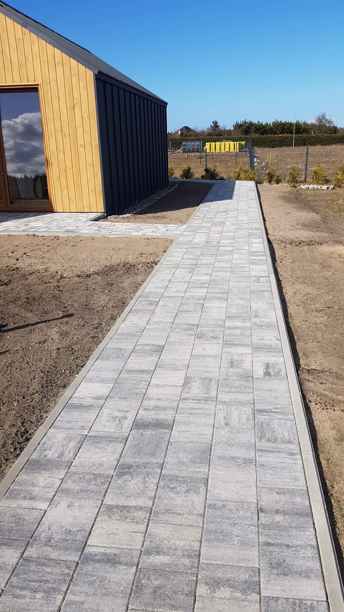 Długa, prosta ścieżka z szarej kostki prowadząca do nowoczesnego domu z drewnianą elewacją i dużym oknem, widok w słoneczny dzień.