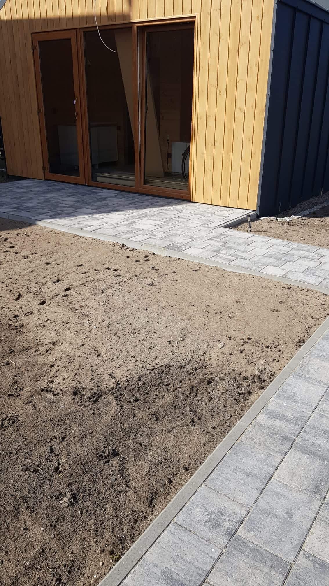 Ułożona szara kostka betonowa przed drewnianym domkiem, z widocznym fragmentem niezagospodarowanego terenu.