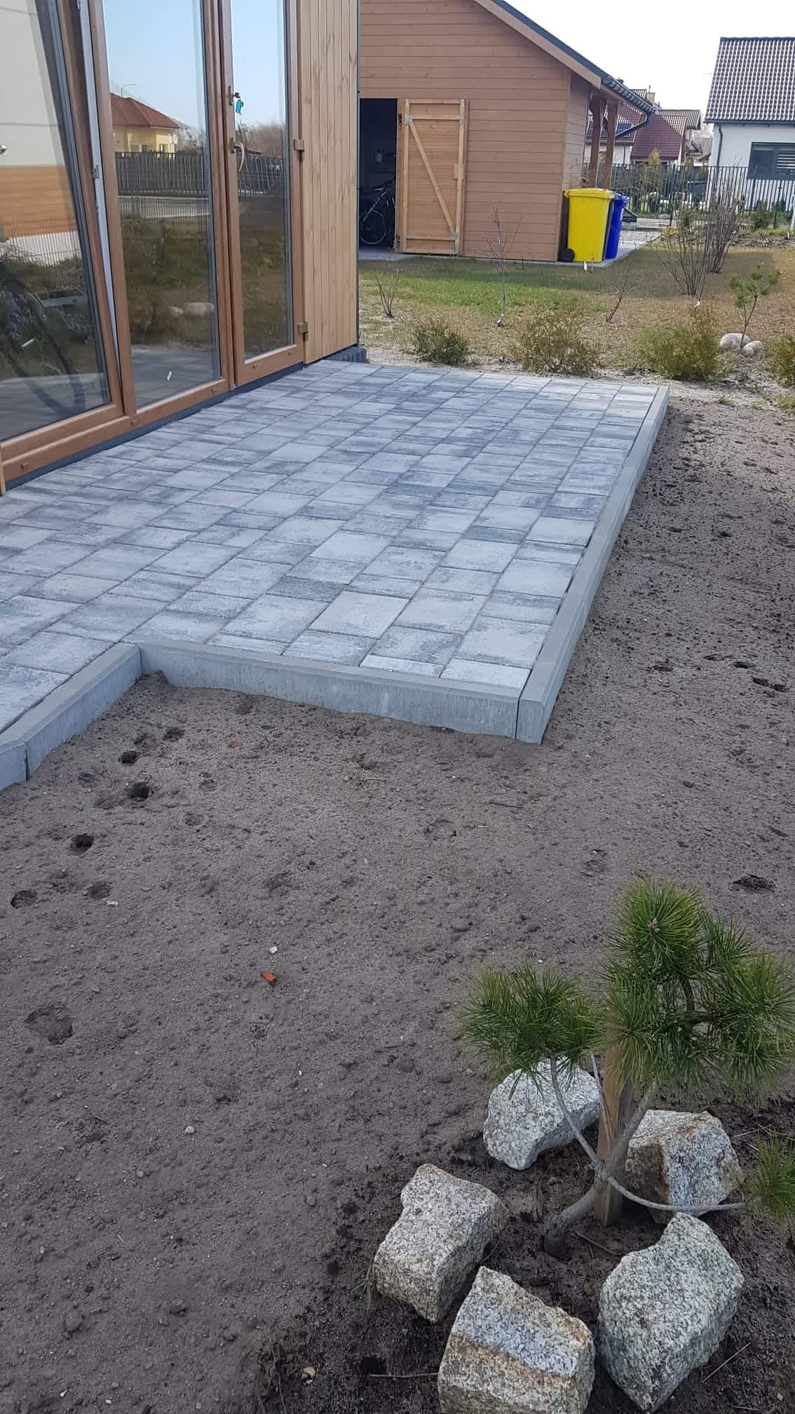 Szara nawierzchnia ułożona z kostki w kwadratowy wzór, obrzeża z szarego betonu, widok na drewniany budynek z przeszklonymi drzwiami i ogród z żółto-niebieskimi koszami na śmieci w tle.