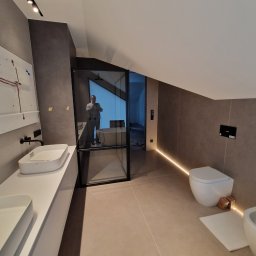 Ferbel - Nowoczesna łazienka na poddaszu w odcieniach szarości z podświetleniem LED podłogi, widoczna kabina prysznicowa ze szklanymi drzwiami i biała armatura: umywalki nablatowe, toaleta oraz bidet.