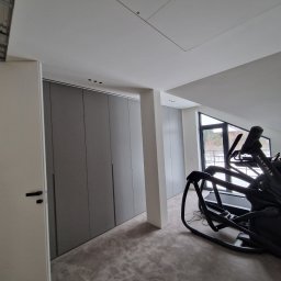 Ferbel - Nowoczesna garderoba z szarymi przesuwnymi drzwiami i czarnymi uchwytami, widoczny sprzęt do ćwiczeń Matrix w pomieszczeniu z oknem i podłogą w odcieniach szarości.