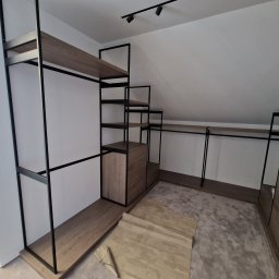 Ferbel - Szafa wnękowa z czarnymi metalowymi ramami i drewnianymi półkami w trakcie montażu na poddaszu, widoczne niedociągnięcia w wykończeniu podłogi i elementy montażowe.