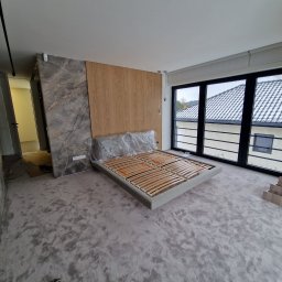 Ferbel - Wnętrze sypialni w trakcie wykańczania: surowa podłoga, łóżko bez materaca zabezpieczone folią, ściana z okładziną imitującą marmur i drewno, duże okno z widokiem na dach.