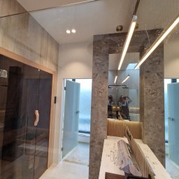 Ferbel - Wnętrze łazienki w trakcie remontu: widoczna sauna z ciemnego drewna, szklana kabina prysznicowa, marmurowe wykończenie ściany z lustrem, oraz przygotowane elementy do montażu armatury łazienkowej.